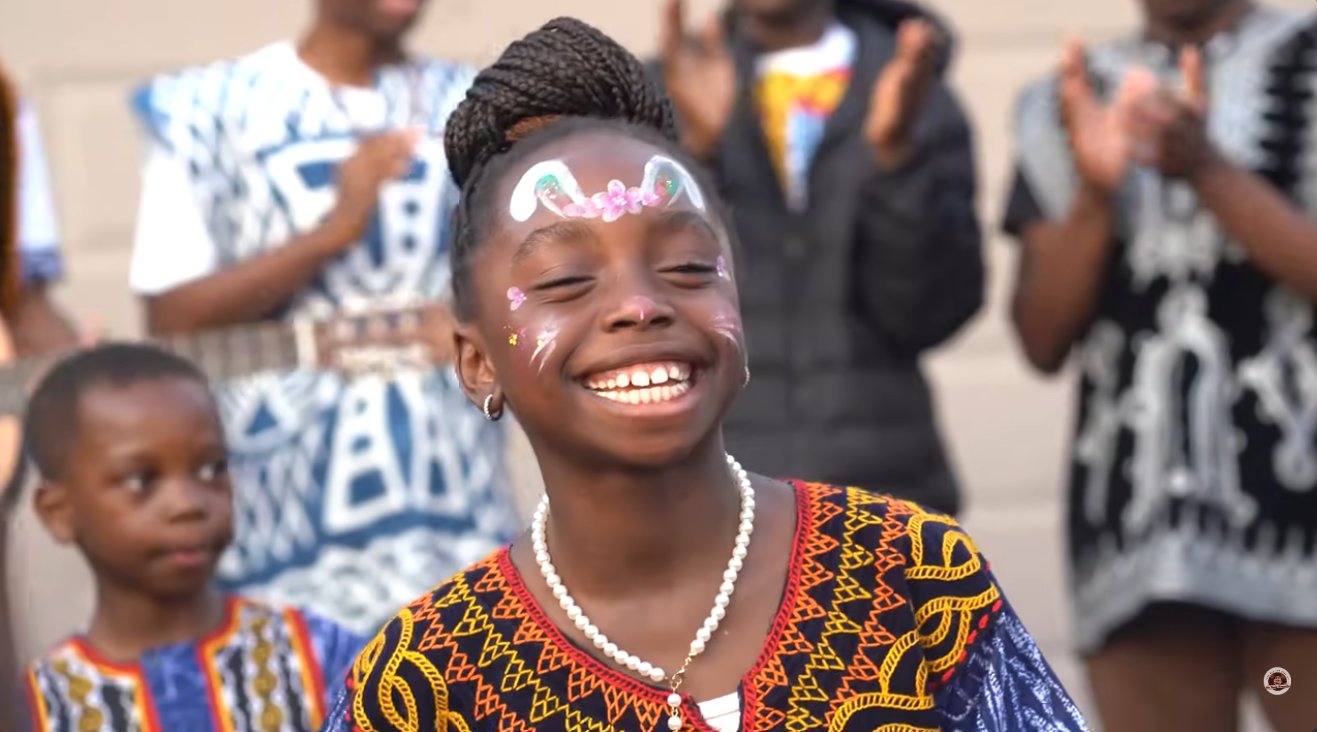 A smiling young girl with face paint and a pearl necklace, wearing a traditional Kom embroidered top, as adults in Kom attire clap behind her