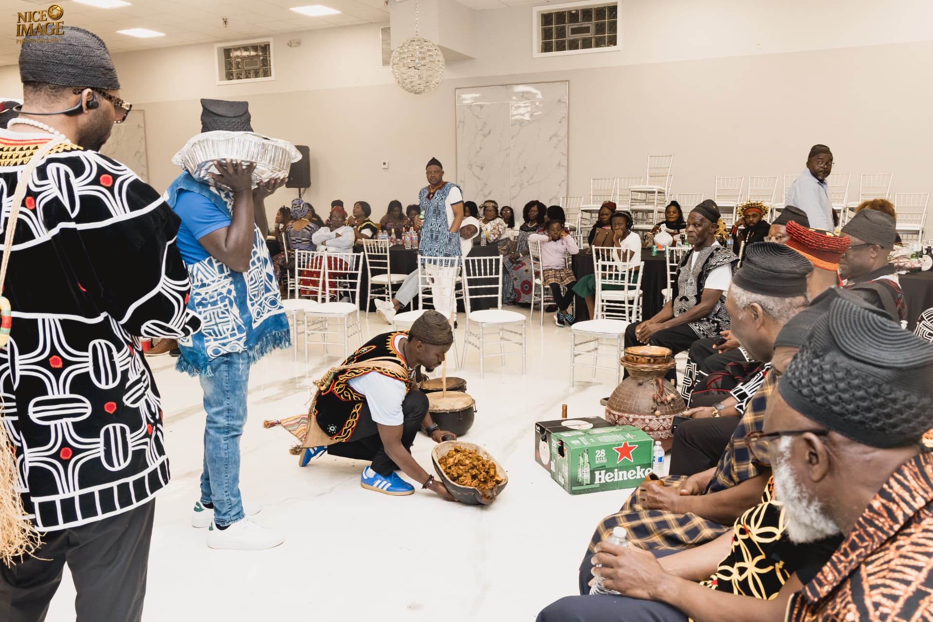 Seated elders in traditional attire observe a ceremonial food presentation accompanied by hand drums and calabash vessels at a BotangRecords cultural gathering.